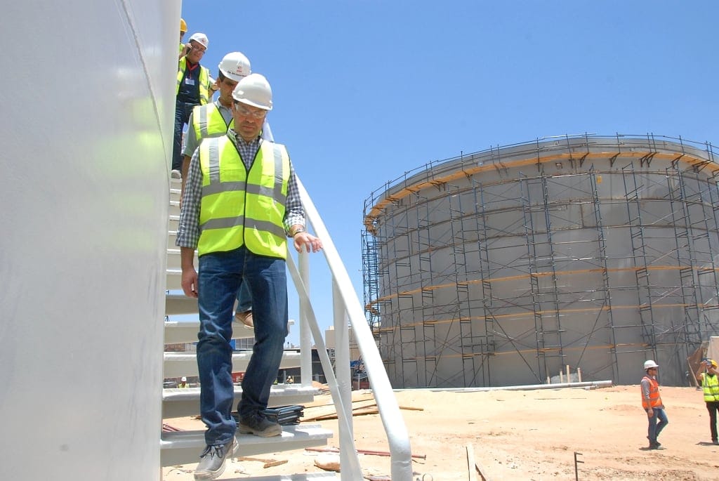 Engineers in high-visibility vests and hard hats descending the stairway of a storage tank, with a scaffolded tank under construction behind