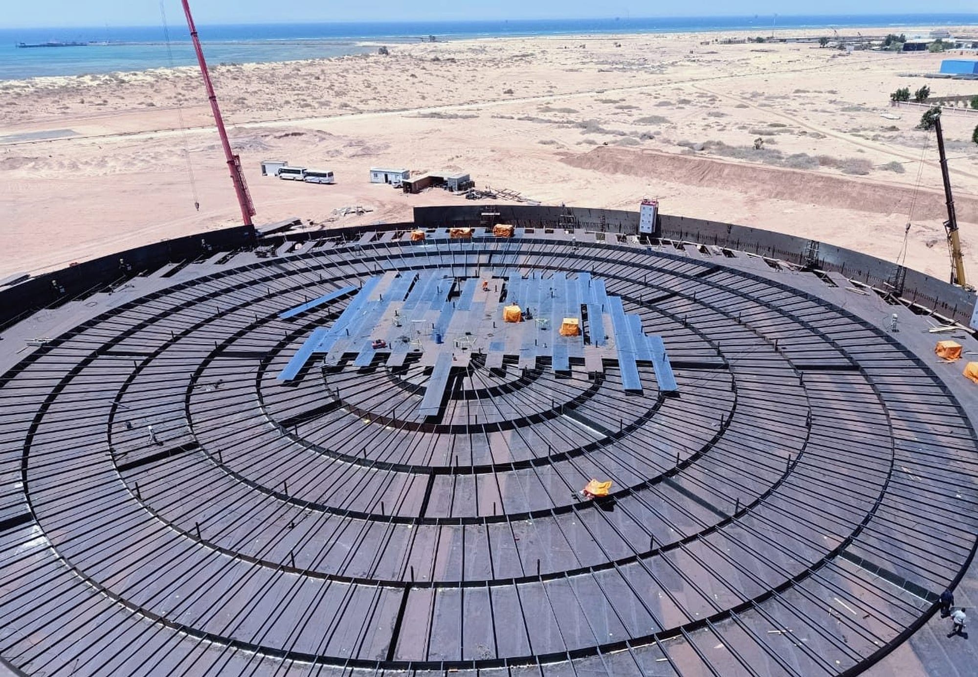 Overhead view of a storage tank under construction, showing radial roof rafters inside the shell and the coastline behind
