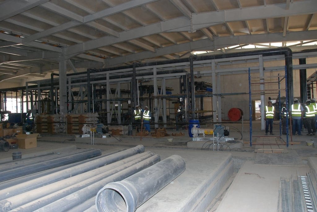 Interior of a plant building during mechanical installation, with large-bore pipe spools laid out and pipe racks and supports overhead
