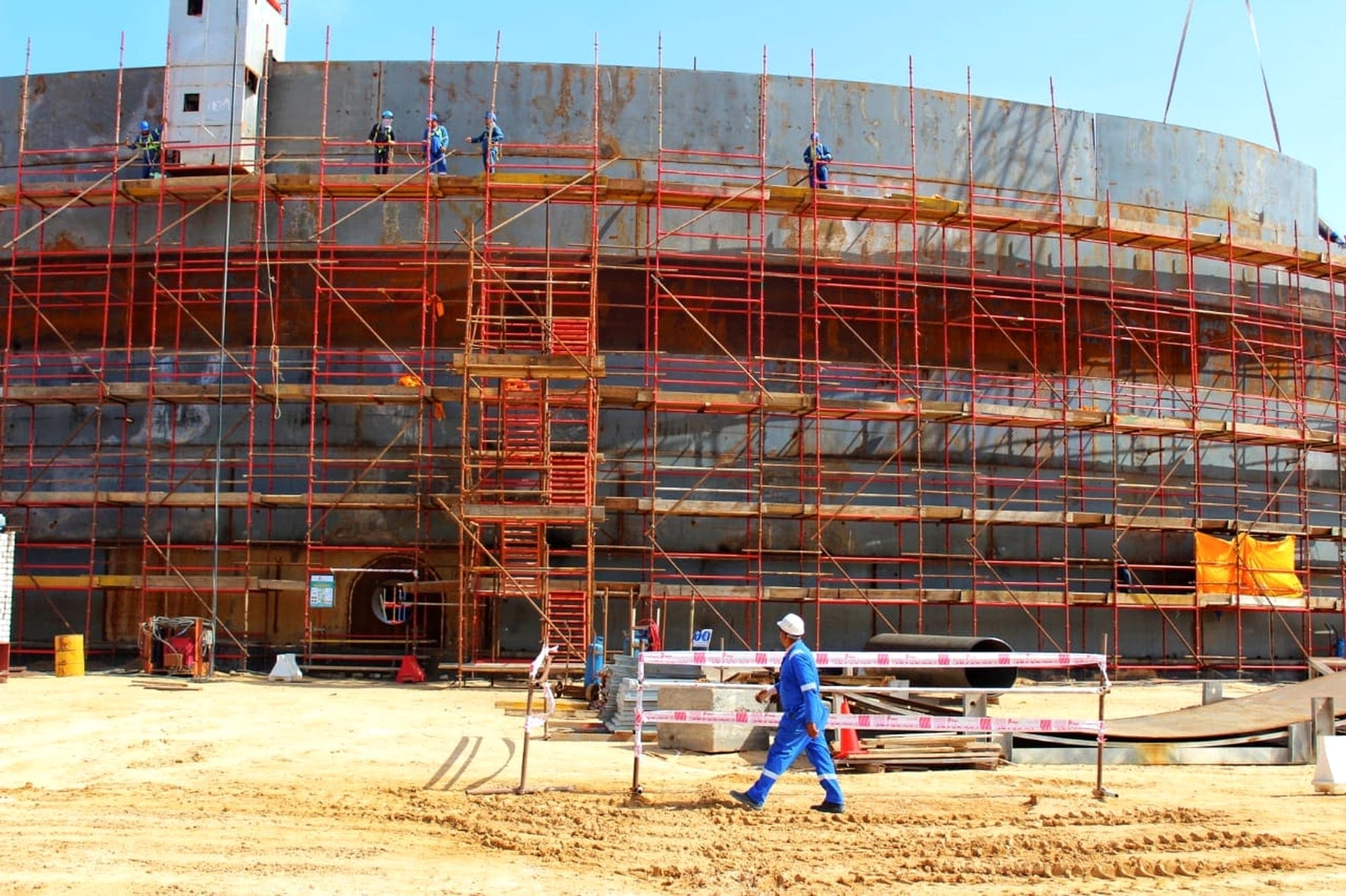 Storage tank shell wrapped in red scaffolding with welders working on the top course